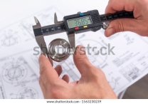 Руки инженера измеряют металлическую деталь цифровым штангенциркулем на фоне технических чертежей. Контроль качества деталей, обрабатываемых на токарном станке.